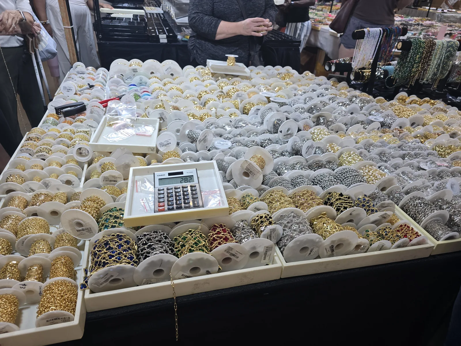 展位长桌铺满上百个金色与银色链条卷,中间一台卡西欧计算器显示 282,旁边摆着镶彩色水钻的花式链条盘,几位买家在挑选