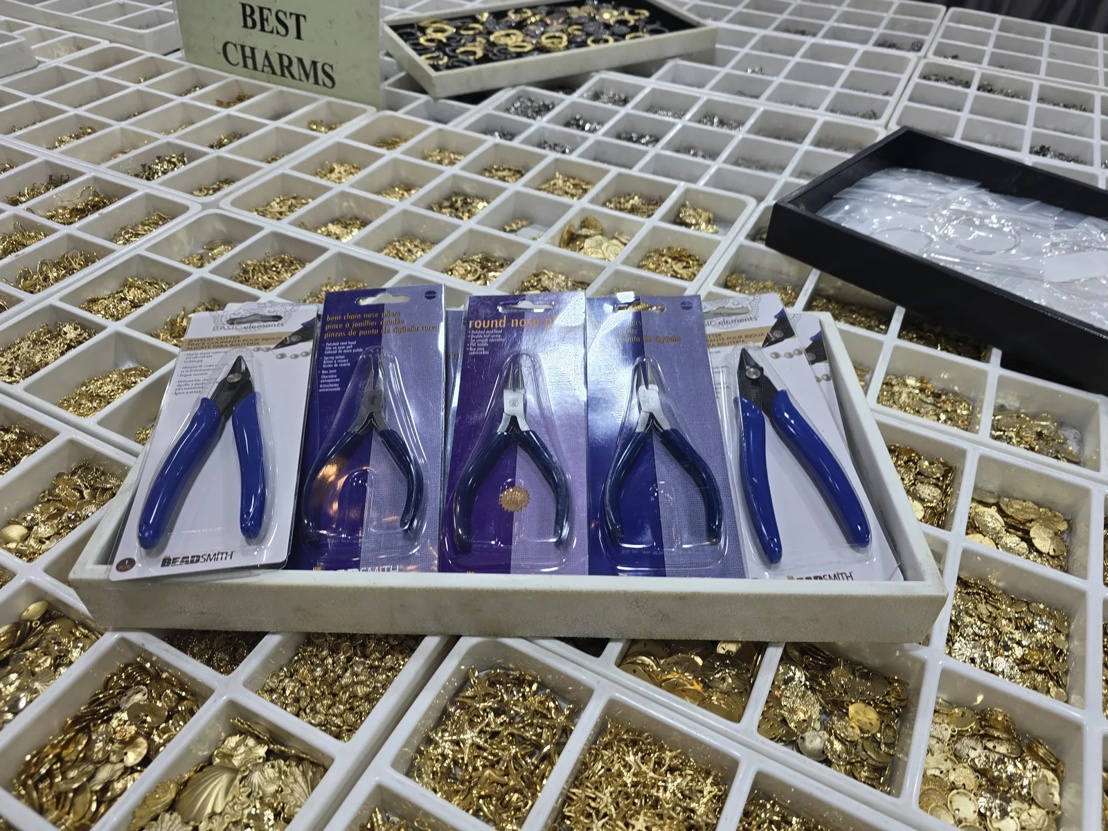 白色分格盘里堆满金色小配件与扣件,中间一盘摆着几把 BeadSmith 品牌的圆嘴钳、剪钳等工具,旁边立牌写着 BEST CHARMS