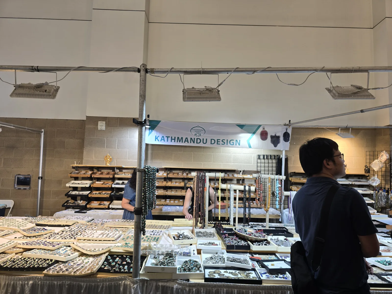 Kathmandu Design 展位横幅下,木质货架陈列银饰与石镶件,前方长桌摆满戒指板与散件,一名观众在旁边观看